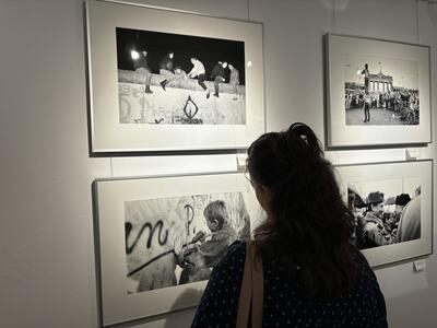 Vier Schwarz-Weiß-Fotografien aus der Zeit des Mauerfalls hängen eingerahmt an einer Wand. Eine Frau steht davor und schaut sie sich an.